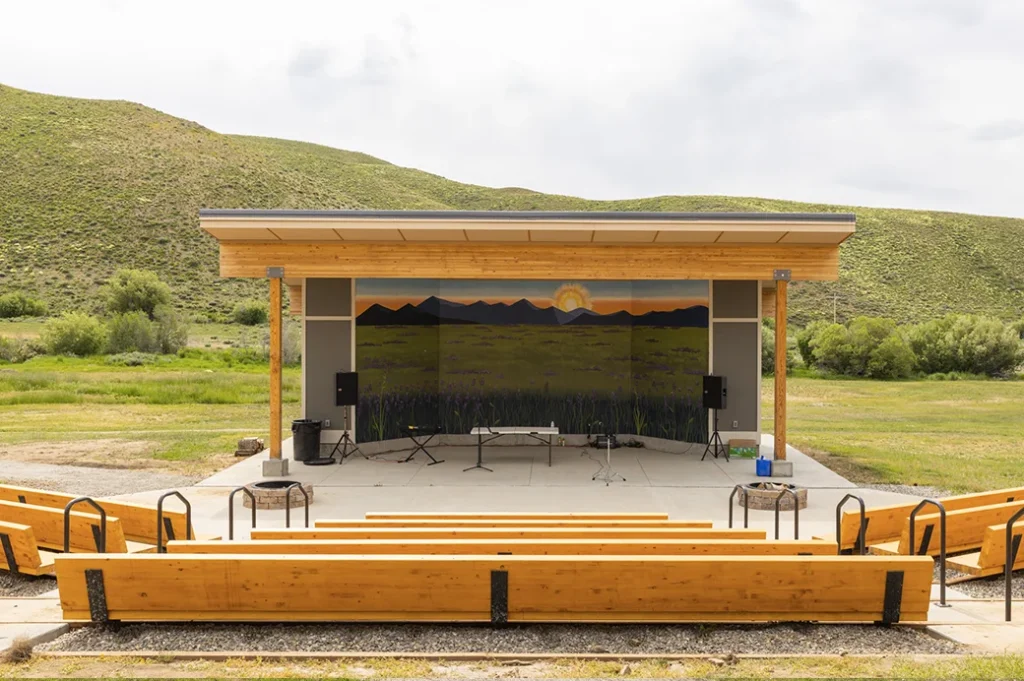 Outdoor amphitheater with wooden benches and a small covered stage featuring a mural of mountains and a sunrise, nestled between grassy hills.”