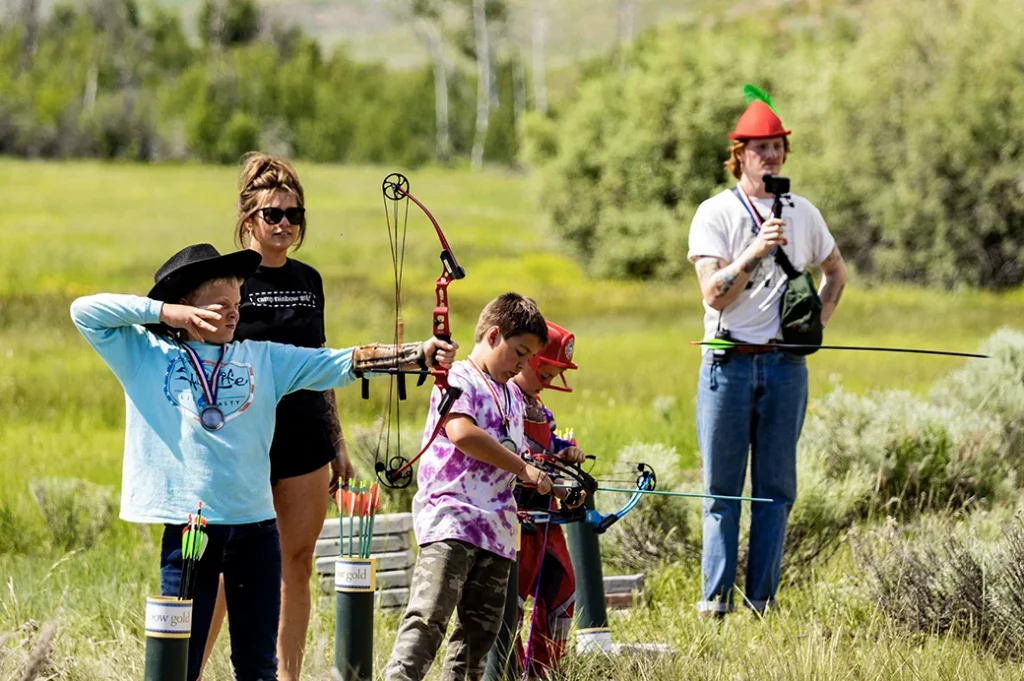 Children practicing archery in a grassy field with bows drawn, supervised by adults, including one dressed playfully in a green hat. Trees and hills line the background.