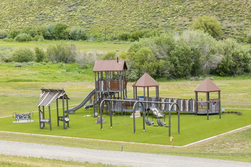 Large outdoor playground with swings, slides, climbing structures, and shaded towers, set on bright green turf amid a scenic grassy landscape.