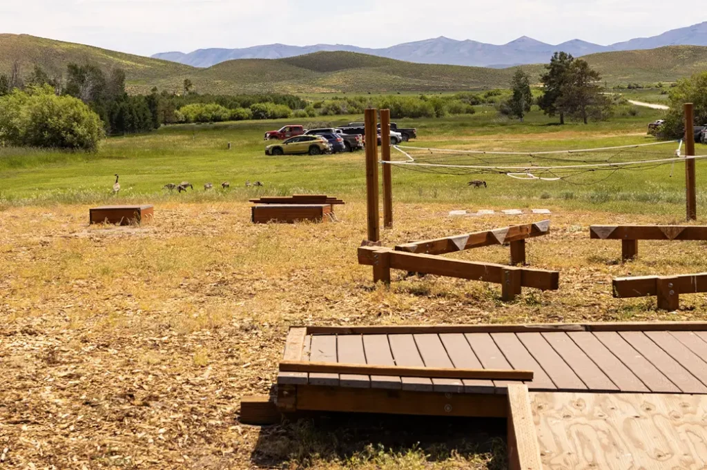 Wooden low-ropes challenge course set in an open field with balance beams and platforms, with cars and geese in the distance and mountains on the horizon.