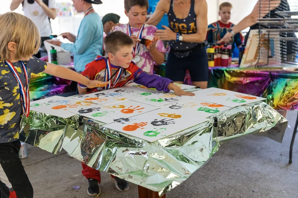 Children wearing medals create colorful handprint art on paper inside a lively craft area decorated with bright foil table covers.