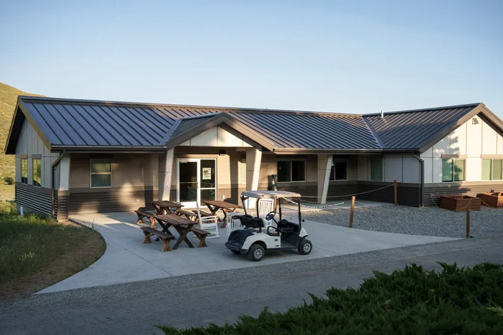 Modern one-story health center with picnic tables and a golf cart parked outside, surrounded by grassy hills and gravel paths.
