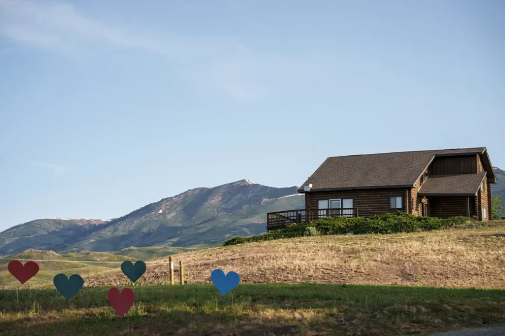 Wooden cabin on a hillside with colorful heart decorations in front and mountains rising in the background under a clear sky.