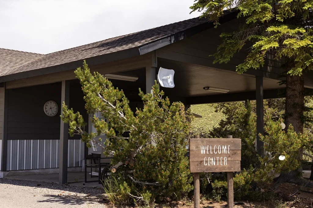 Small gray building with a wooden sign reading ‘Welcome Center,’ surrounded by trees and set against a sunny hillside.